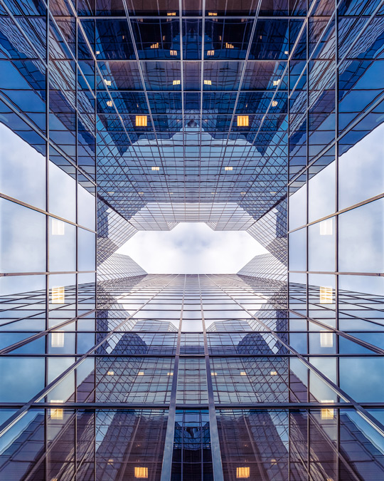An upward looking view of towering glass office buildings creating a symmetrical, geometric pattern against the sky.