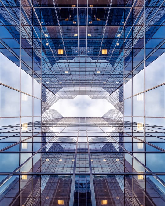 An upward looking view of towering glass office buildings creating a symmetrical, geometric pattern against the sky.