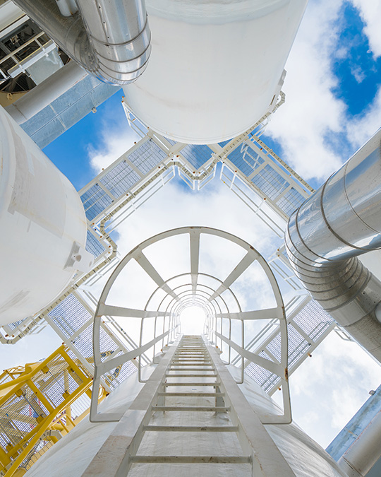 An upward looking view through a caged industrial ladder surrounded by pipes and tanks against a blue, cloudy sky.